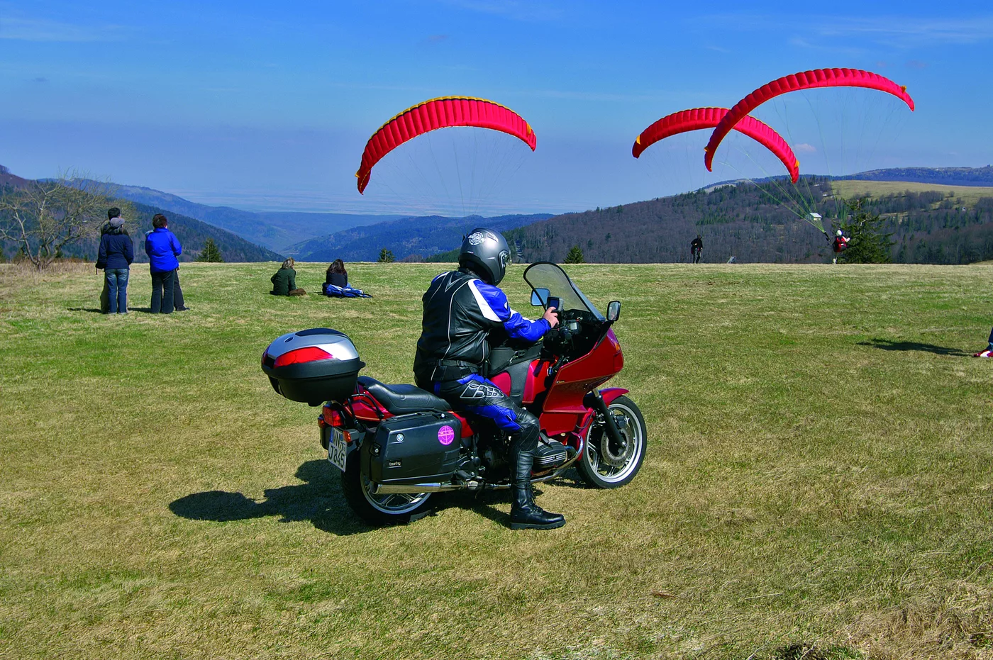 Gleitschirme über der Bergwiese an der Route des Crêtes, davor ein Motorrad Gleitschirme über der Bergwiese an der Route des Crêtes, davor ein Motorrad
